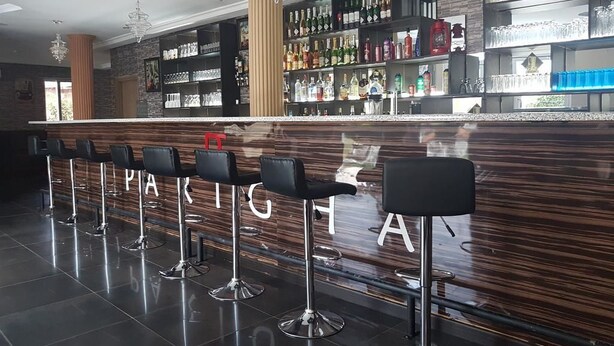 A bar area with a wooden counter, multiple bar stools, and shelves stocked with bottles and glasses. - Parigha Hotel (Accra)