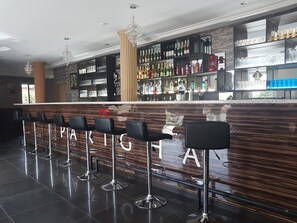 A bar area with a wooden counter, multiple bar stools, and shelves stocked with bottles and glasses. - Parigha Hotel (Accra)