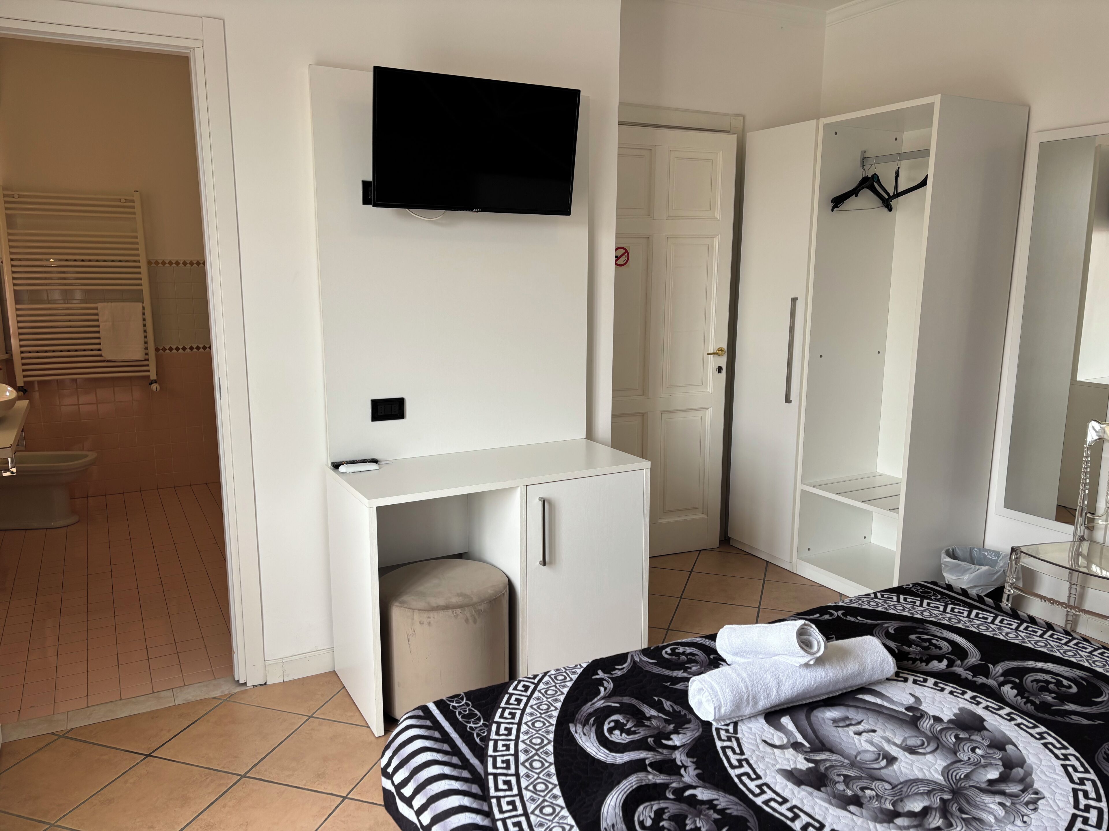 A hotel room with a bed, a TV mounted on the wall, a wardrobe, a mirror, and a bathroom visible through an open door.