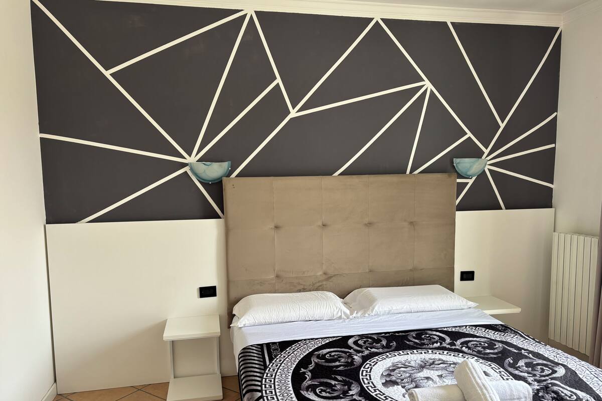 Un dormitorio con un cabecero estampado con motivos geométricos, una cama con colcha estampada, dos mesitas de noche y una lámpara de pared.