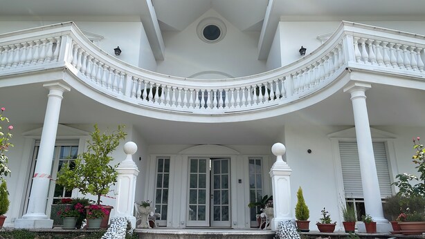 Imponente entrada con un balcón curvo, columnas blancas y una puerta central con paneles de vidrio.