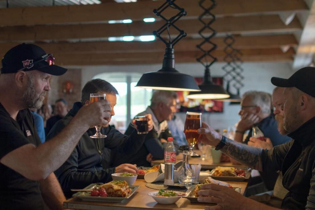 Eine Gruppe von Menschen sitzt an einem Tisch mit Speisen und Getränken in einem Raum mit hängenden Lampen und Holzbalken.