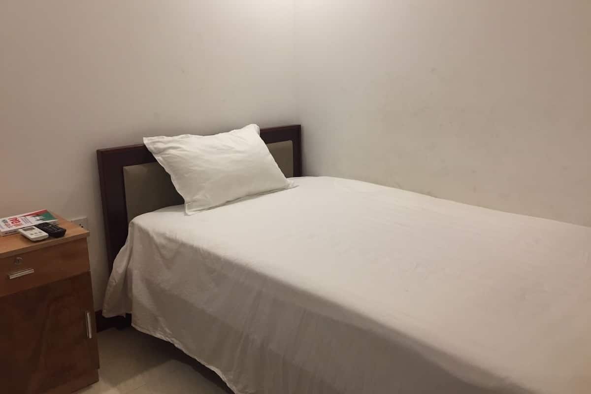 A single bed with a white mattress and pillow, a wooden nightstand with a remote control, and a small drawer.