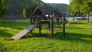 A playground with a wooden structure, including a slide and climbing elements, set in a grassy area with trees and a hill in the background. - Hotel Garni Reis (Treis-Karden)
