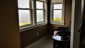 Ein Eckzimmer mit drei Fenstern und Blick auf ein Gewässer, einem kleinen Tisch und einem Stuhl.