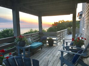 A wooden deck with a bench and chairs, overlooking a body of water at sunset.