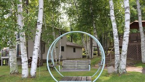Un ensemble de balançoires en bois avec une structure métallique circulaire, entouré de bouleaux et avec une maison en arrière-plan.