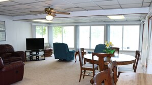 Un salon spacieux doté d’un ventilateur de plafond, d’un téléviseur à écran plat, d’un canapé en cuir, d’une table à manger avec des chaises et de grandes fenêtres offrant une vue sur la verdure.