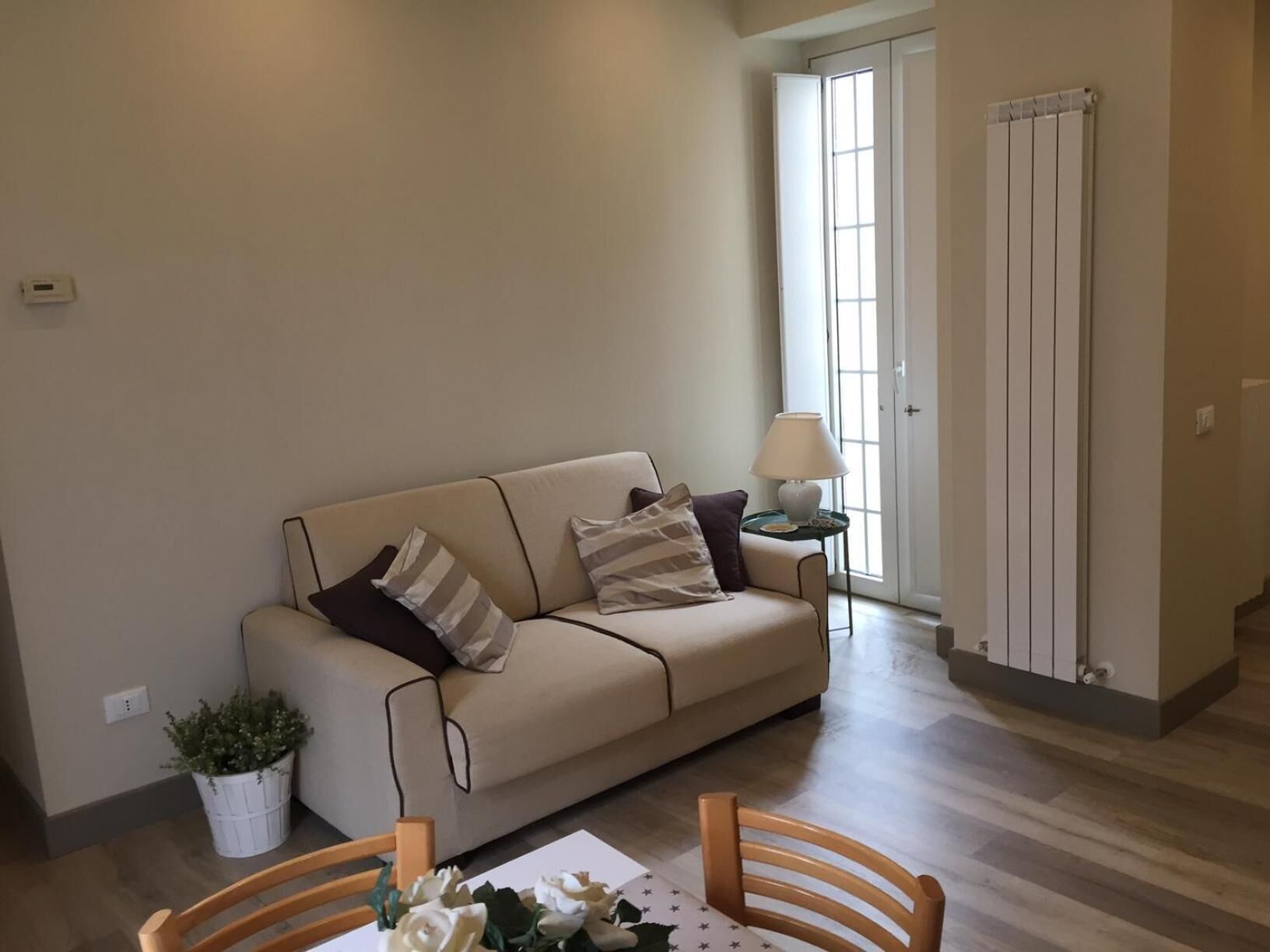 Un salon avec un canapé beige, deux chaises en bois, une petite table avec une lampe, une plante en pot et un radiateur.