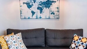 Ein modernes Wohnzimmer mit einem dunkelgrauen Sofa, gemusterten Kissen, einem runden Holztisch mit Obstschale und einer Weltkarte als Wandkunst.
