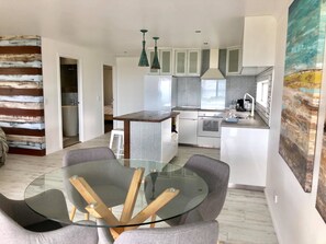 A modern kitchen with a central island, white cabinetry, and a dining area with a glass table and grey chairs. - Sky Loft Coffs · Sky Loft On Bruxner - Korora (Korora)