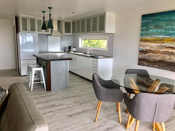 A modern kitchen with white cabinets, a central island, and a dining area with a glass table and grey chairs. - Sky Loft Coffs · Sky Loft On Bruxner - Korora (Korora)
