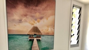 Une chambre à coucher avec un lit, des oreillers, une table de chevet, une lampe de chevet, une fenêtre avec des stores et un grand tableau représentant une scène de plage.