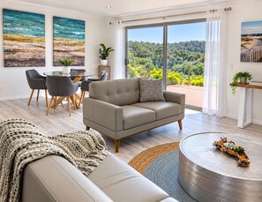 Uma sala de estar moderna com um sofá, um espaço de refeições com cadeiras e vista para uma paisagem arborizada através de grandes janelas.