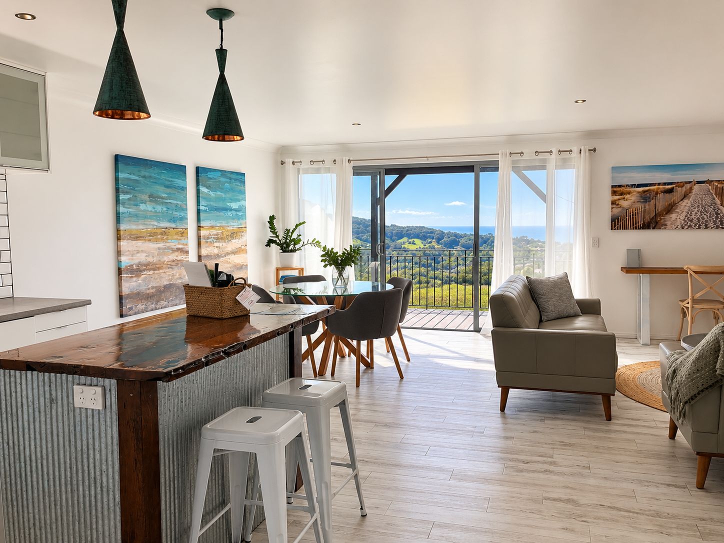 A modern kitchen with a central island, dining area with chairs, a sofa, and a balcony with a view.