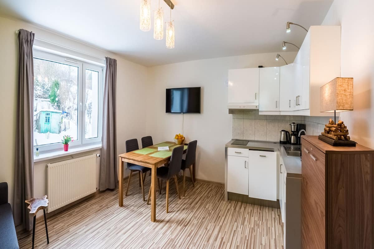 Une cuisine moderne avec des meubles blancs, une table à manger avec des chaises, un téléviseur fixé au mur et une fenêtre avec des rideaux.