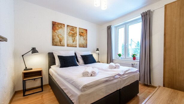 A neatly made bed with white linens and rolled towels, a bedside table with a lamp, a window with curtains, and a small potted plant on the windowsill. - RentPlanet - Apartament Bronka Czecha (Swieradow-Zdroj)
