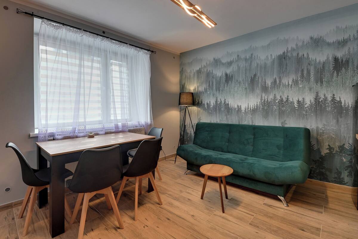 Une pièce comprenant une table à manger, des chaises, un canapé vert, une petite table ronde et une fresque représentant une forêt sur le mur.