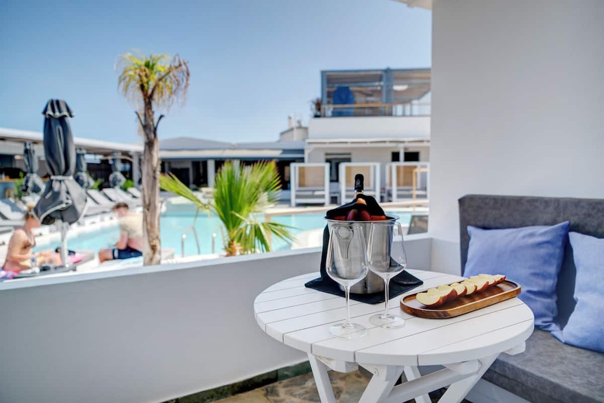 A white table with a bottle of champagne, two glasses, and a wooden tray with sliced apples on a balcony overlooking a pool area.