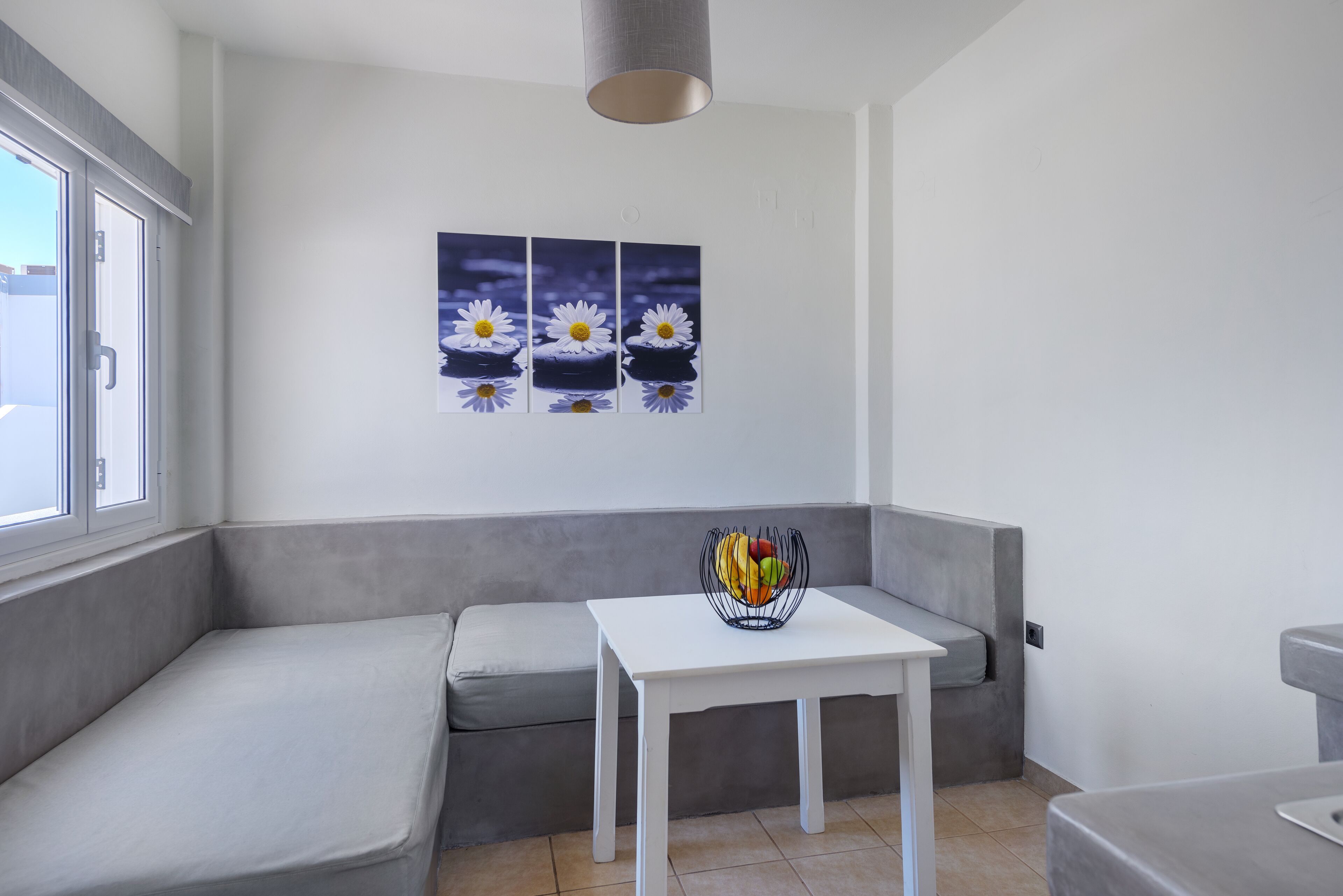 Una habitación moderna con un sofá gris, una mesa blanca y un frutero. Hay tres cuadros de flores en la pared.