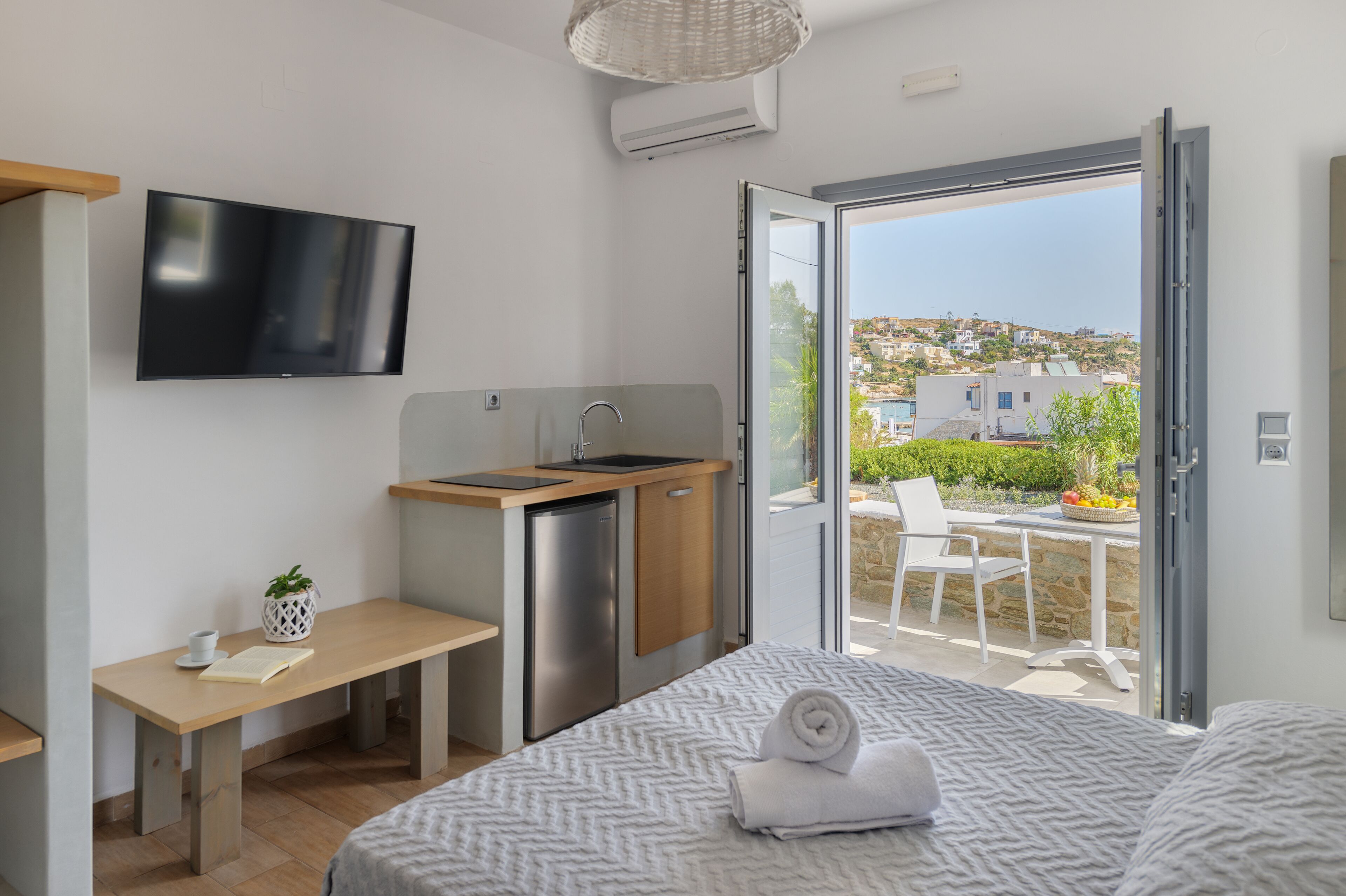A modern hotel room with a bed, a small kitchenette, a TV, and a balcony with a view of the city.