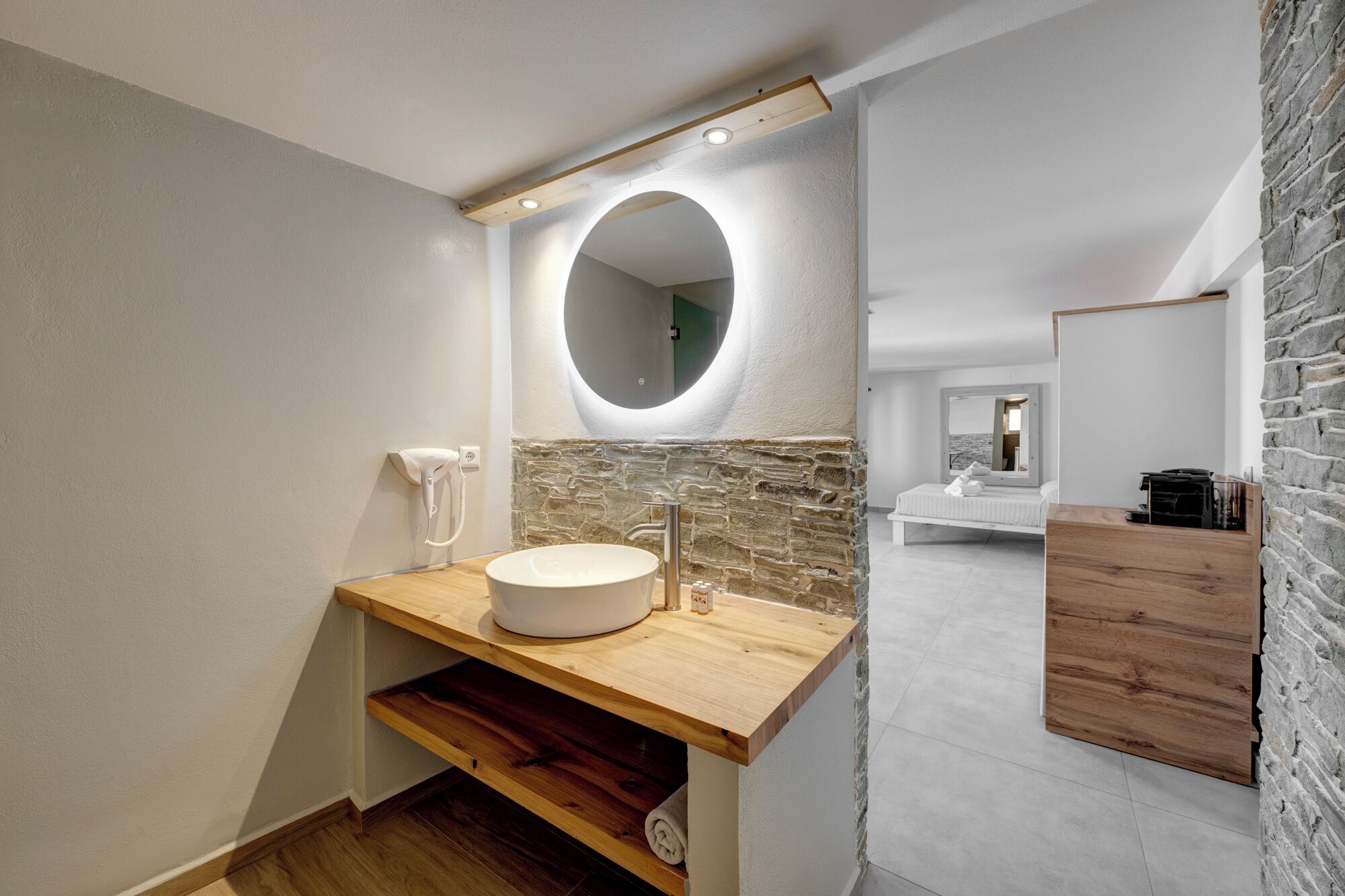 A modern bathroom with a stone accent wall, a wooden vanity, a round mirror, and a view of the bedroom.