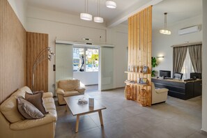 A modern hotel lobby with a sofa, a coffee table, a reception desk, and a view of the outside. - Aegean Paradiso Vacation Club Adults Only (Syros)