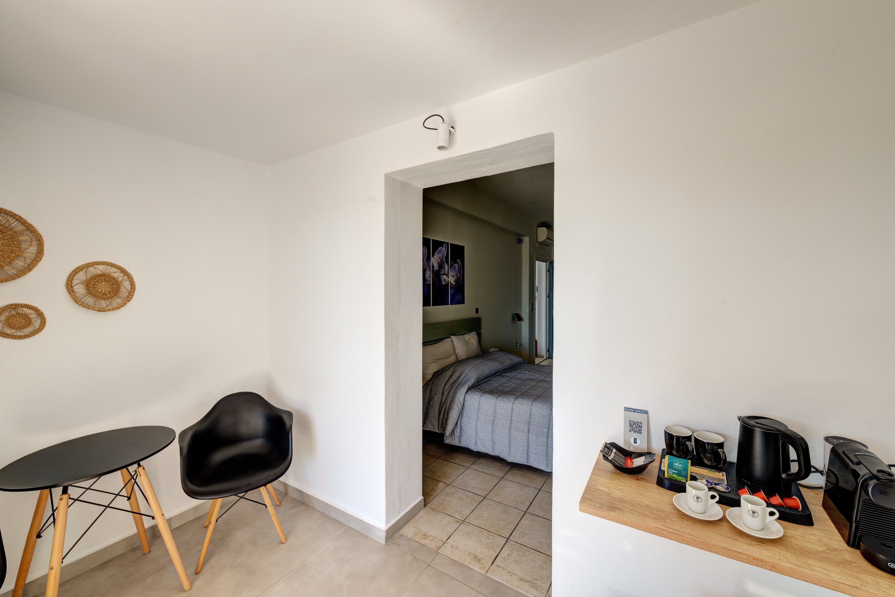 A compact room with a bed, a small table, a chair, and a kitchenette area with a coffee machine and cups.