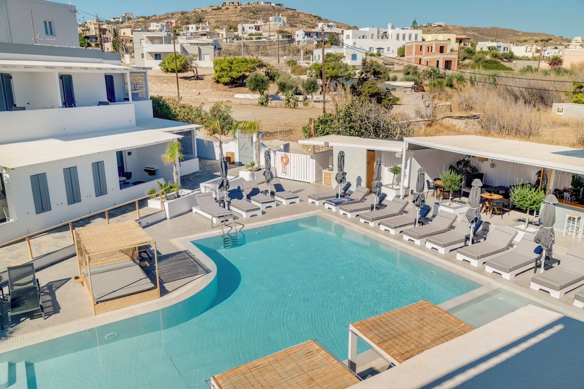 A modern pool area with lounge chairs and a clear blue pool, surrounded by white buildings and a hillside in the background.
