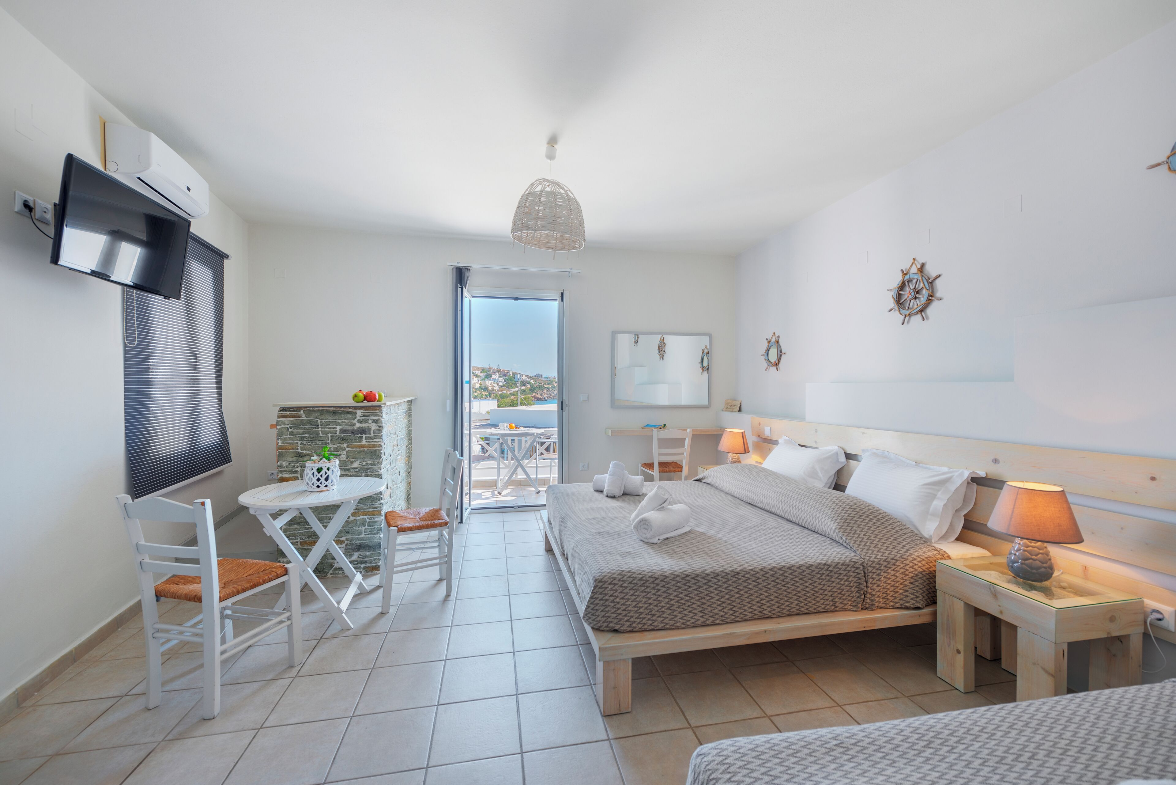 A bedroom with a bed, bedside tables, a TV, a small table with chairs, and a view of the outside.