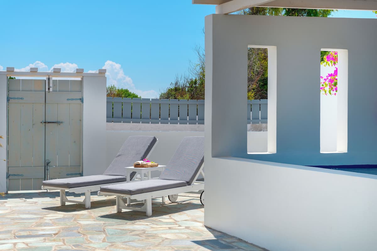 A poolside area with two lounge chairs and a small table, a white wall with a window, and a gate in the background.