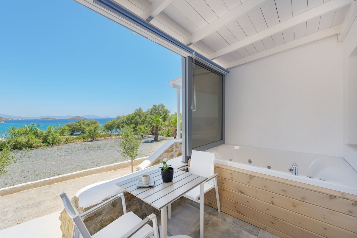 A balcony with a view of the sea, a wooden table, a white chair, and a bathtub.