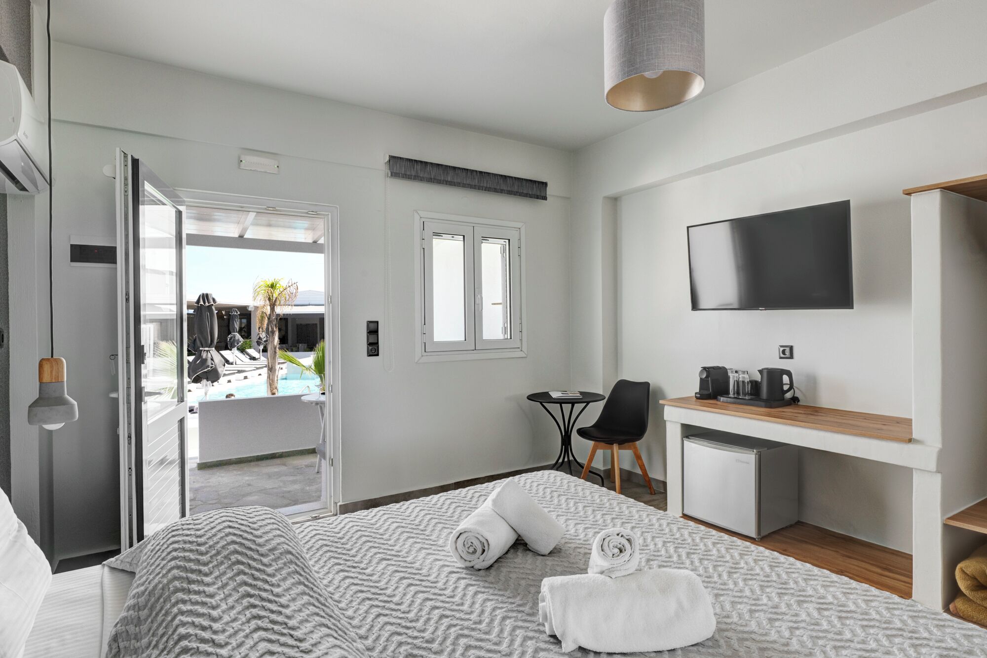 Une chambre d’hôtel moderne équipée d’un lit, d’un bureau avec une télévision, d’une petite table et d’une chaise. Une fenêtre offre une vue sur une piscine et des palmiers.