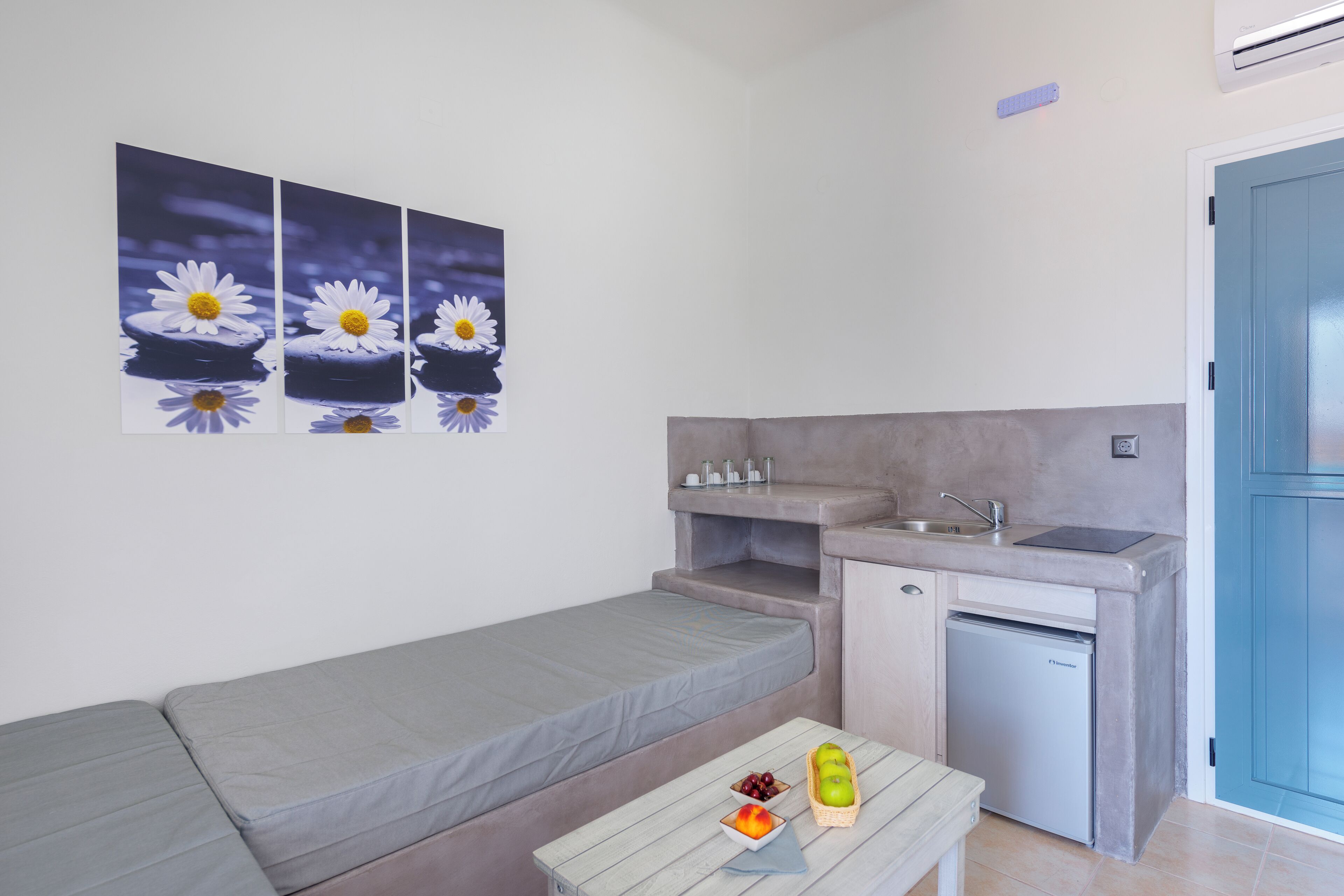 Habitación compacta con sofá gris, una kitchenette pequeña con lavamanos y un mini refrigerador, y una mesa con frutas y un tazón.