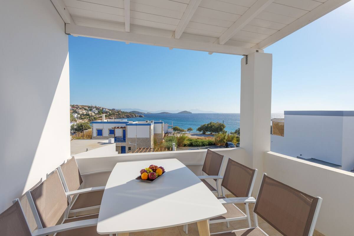 A balcony with a white table and chairs, a fruit tray, and a view of the sea and buildings.