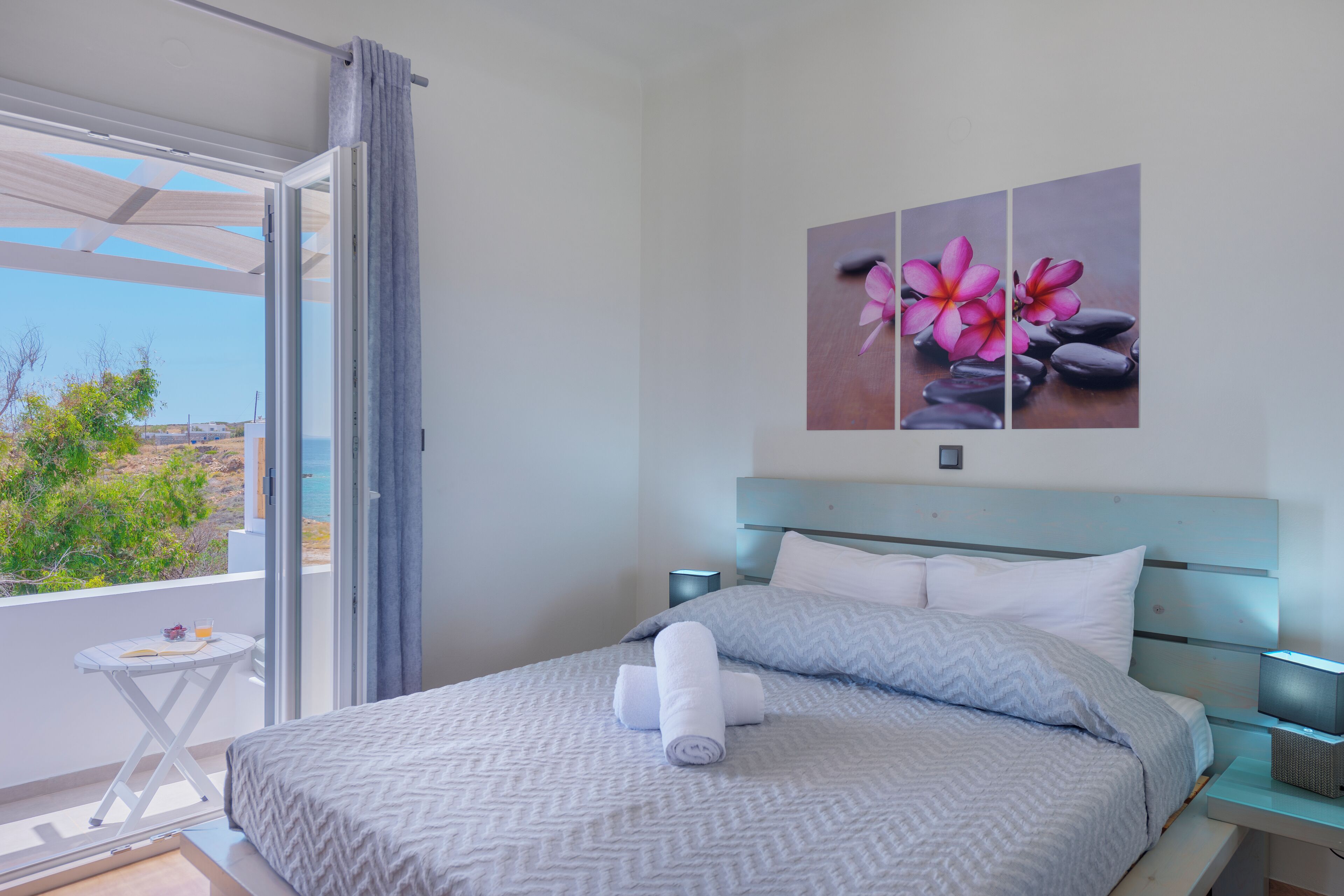 A bedroom with a bed, pillows, a nightstand, and a view of the sea through a sliding door.
