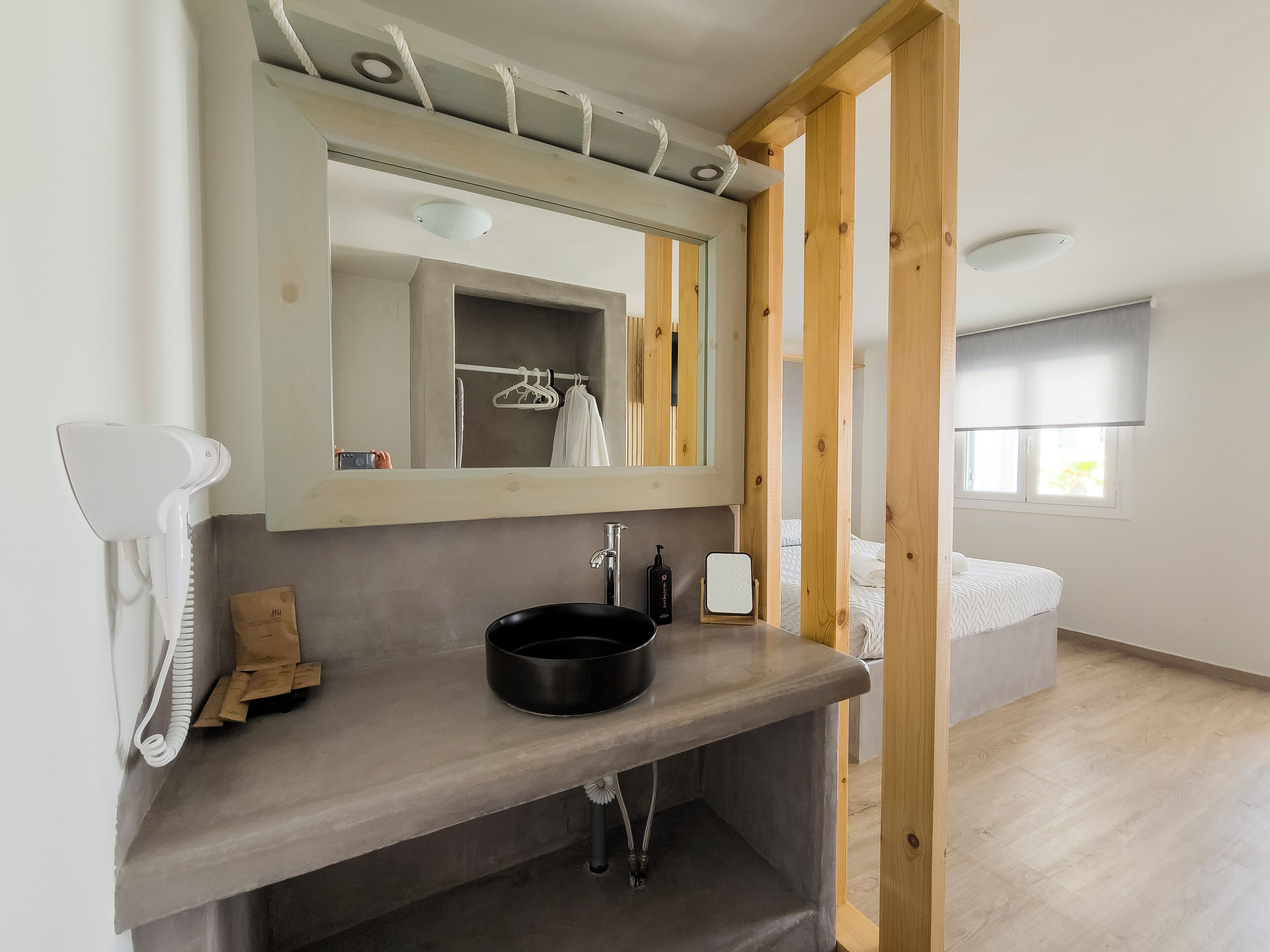 Um banheiro moderno com pia cinza, um espelho e um secador de cabelo. Há uma janela com vista para uma cama e uma persiana.