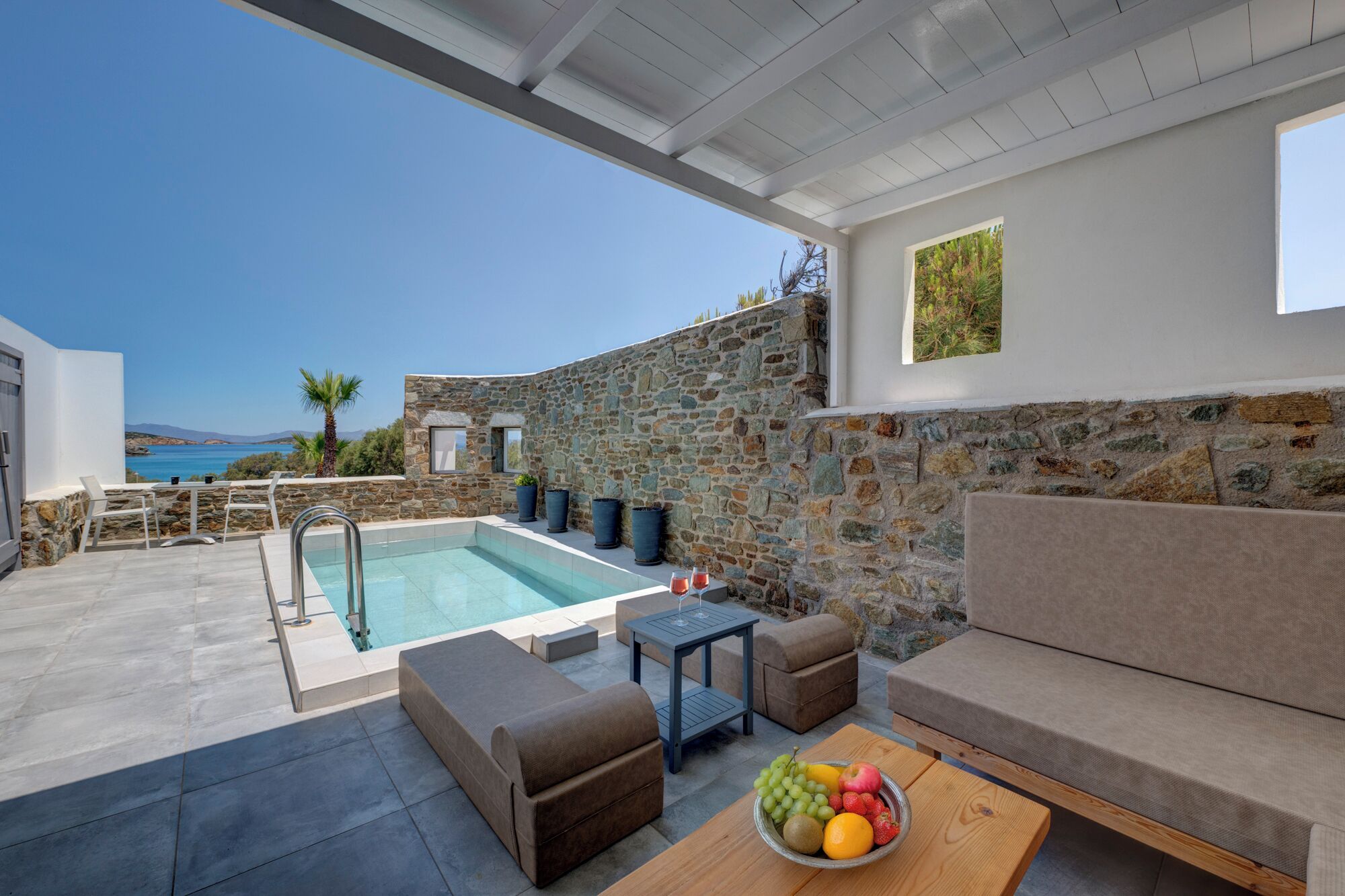Zona junto a la piscina con pared de piedra, mobiliario exterior y vista al mar.