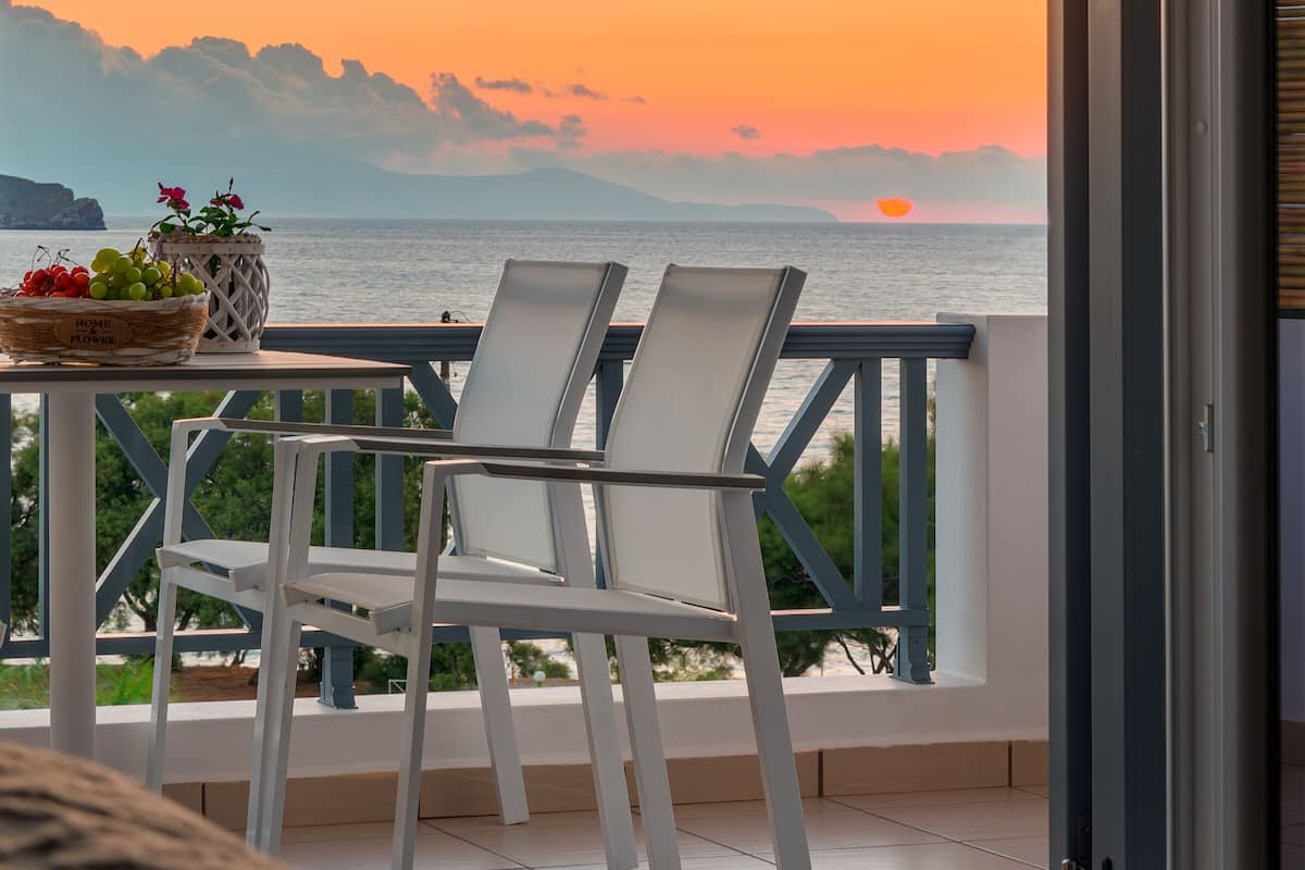 A balcony with a view of the sunset over the sea, a table with a basket of fruit, and two white chairs.