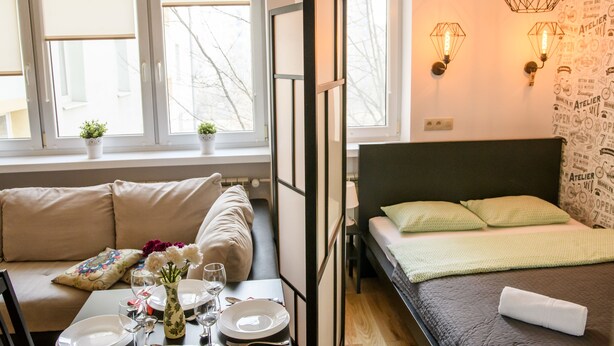 A room with a bed, a sofa, a dining table set with plates, glasses, and a vase of flowers. There are windows with blinds, a wall with light fixtures, and a small plant on the windowsill. - Chmielna Superior (Warsaw)
