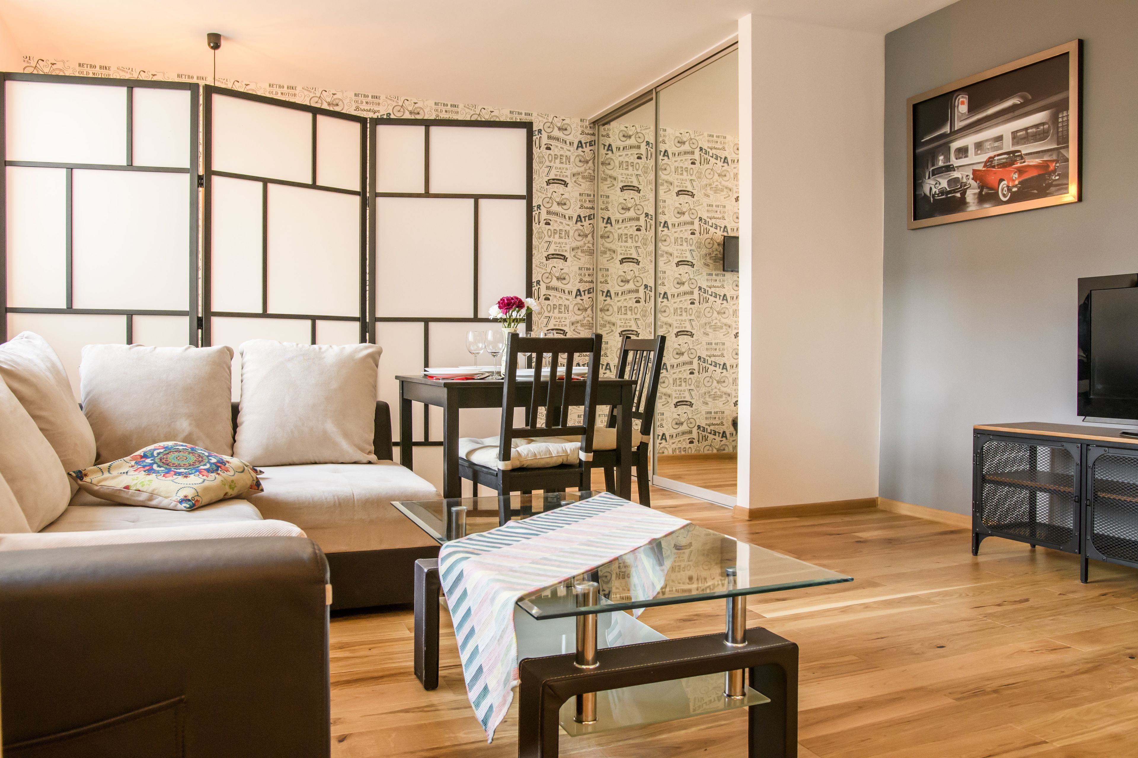 A modern living room with a sofa, a glass coffee table, a dining table with chairs, a TV stand, and a decorative wall with a vintage car print.