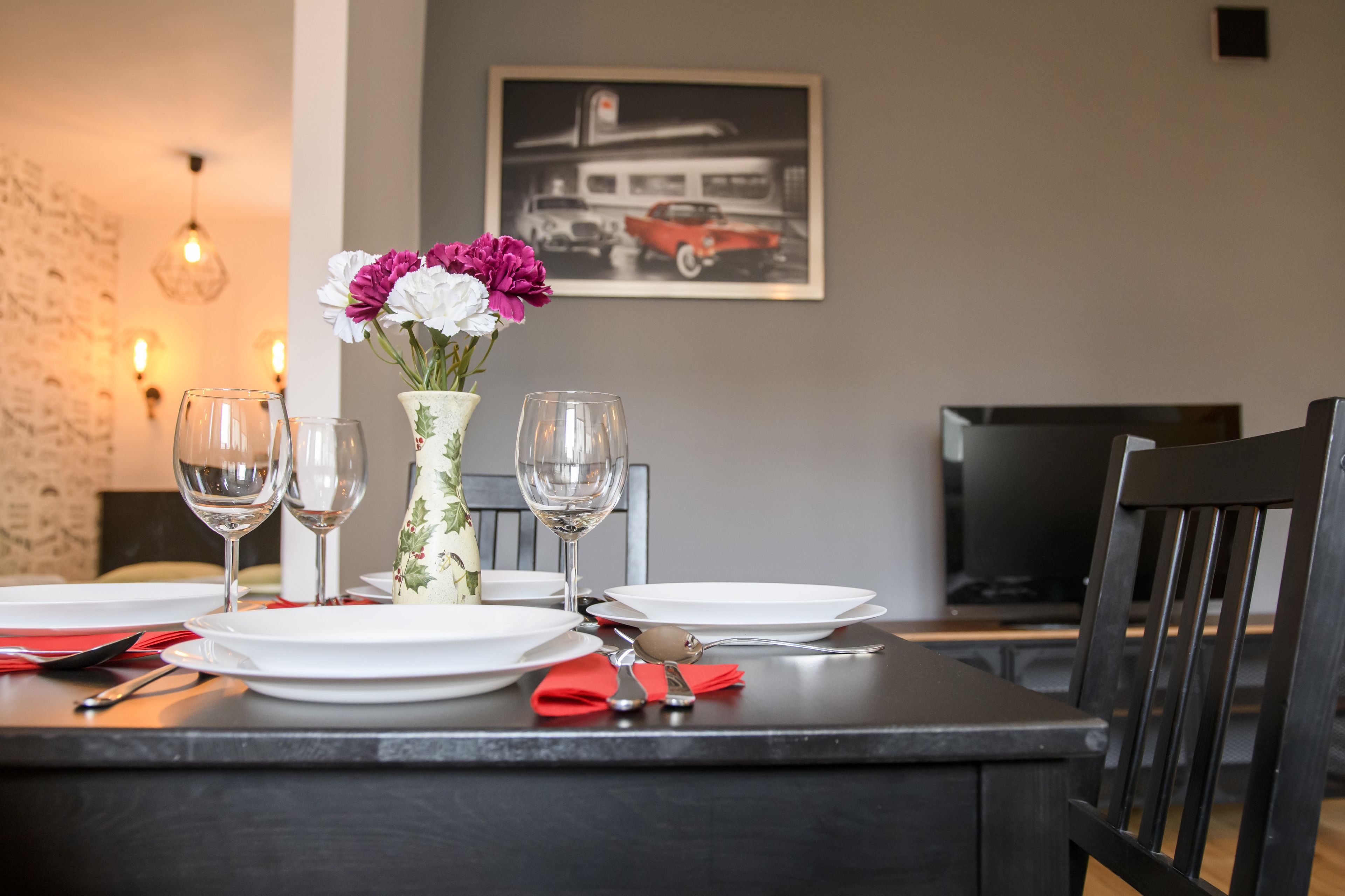 A dining table set with plates, glasses, and cutlery, a vase with flowers, and a framed picture on the wall.