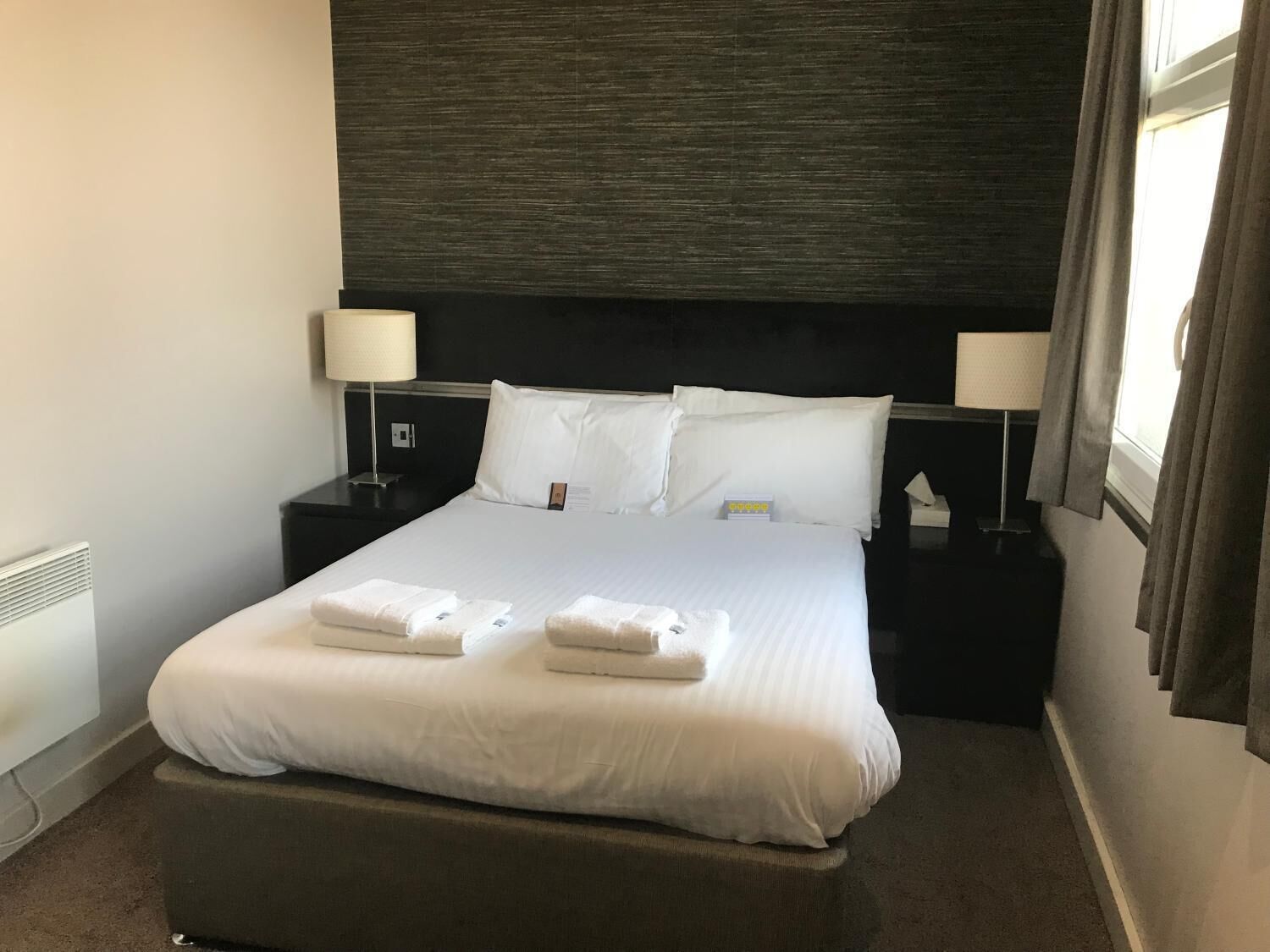 A neatly made bed with white linens and two towels, a dark headboard, two bedside tables with lamps, and a window with curtains.