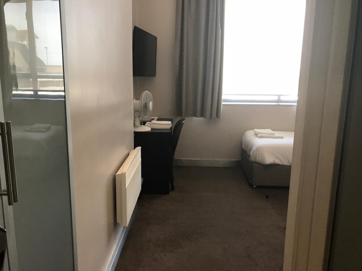 Une chambre d’hôtel comprenant un lit, un bureau avec une chaise, un téléviseur à écran plat fixé au mur, une fenêtre avec des rideaux et un radiateur.