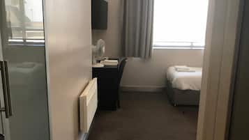 Habitación de hotel con cama, escritorio con silla, televisión de pantalla plana en la pared, ventana con cortinas y radiador.