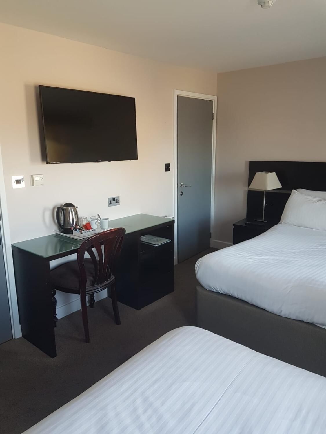Et hotelværelse med en seng, et skrivebord med stol, et fjernsyn, en dør og et natbord med lampe.