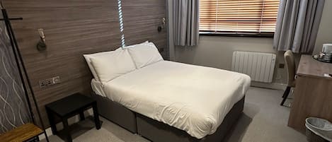 Habitación de hotel moderna con una cama grande, una mesita de noche, un taburete pequeño, un radiador y una ventana con persianas.