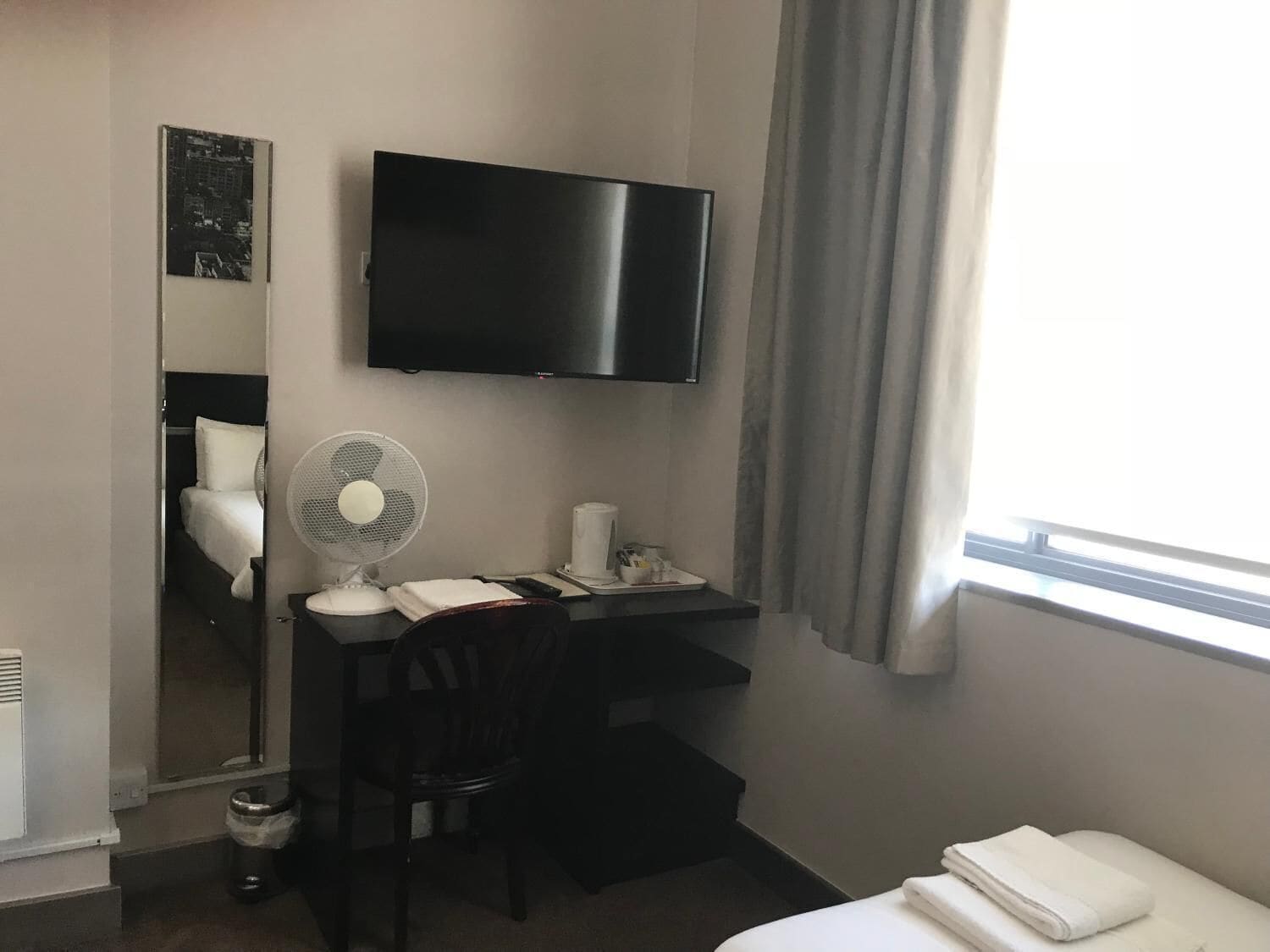 A hotel room with a bed, a desk with a chair, a TV, a fan, and a window with curtains.