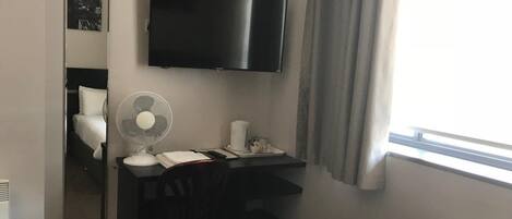 A hotel room with a bed, a desk with a chair, a TV, a fan, and a window with curtains.