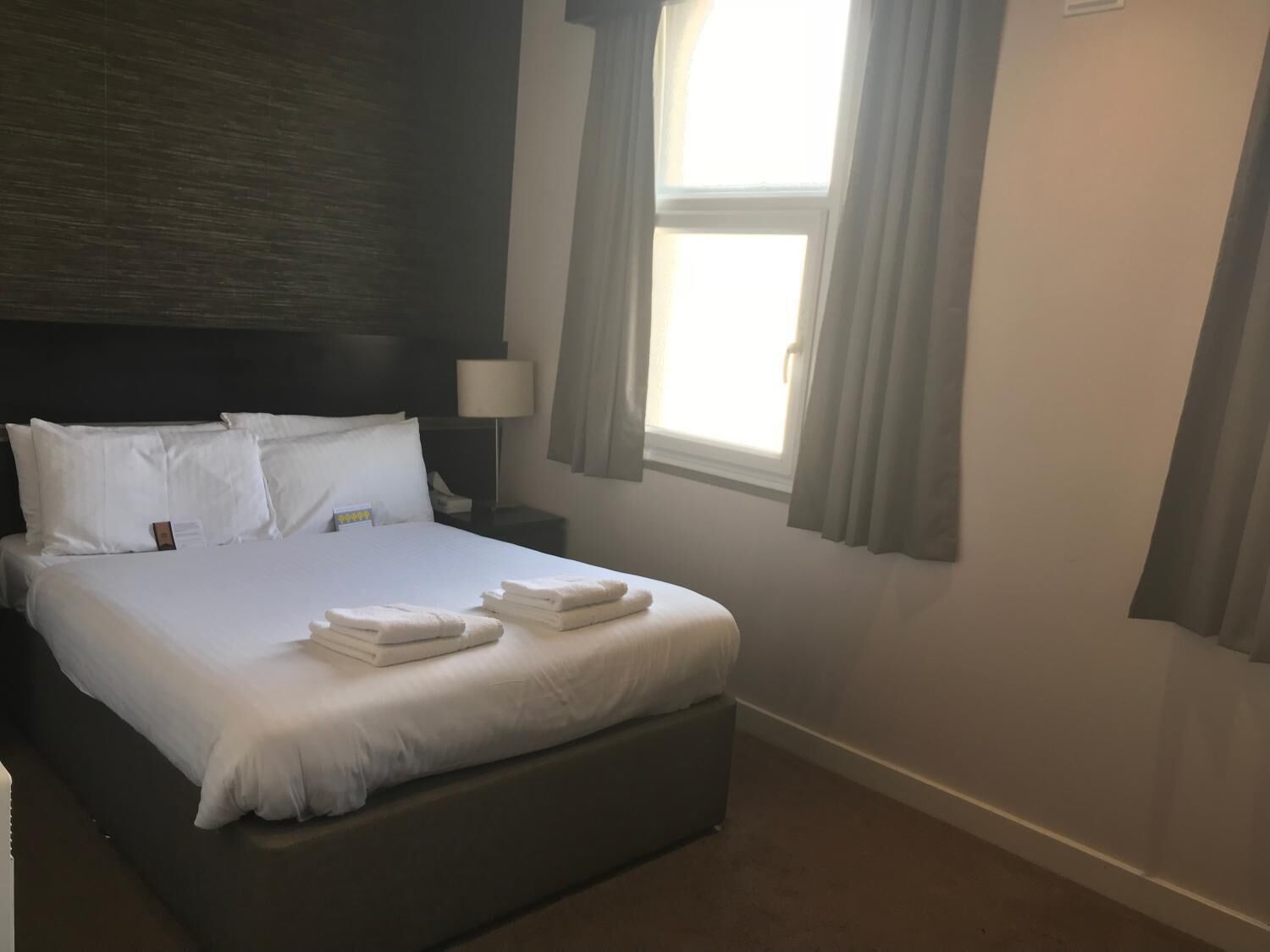 A neatly made bed with white linens and towels, a dark headboard, a bedside lamp, and a window with curtains.